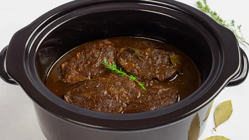 Vier stukjes draadjesvlees in de slowcooker met een takje groen en een laurierblaadje.