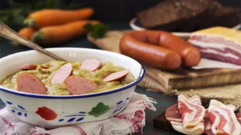 Erwtensoep recept van oma in een ouderwets kommetje met rookworst en plakjes spek ernaast.