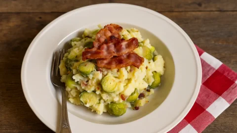Een bord spruitjesstamppot met spek en mosterdsaus op een geblokte theedoek.